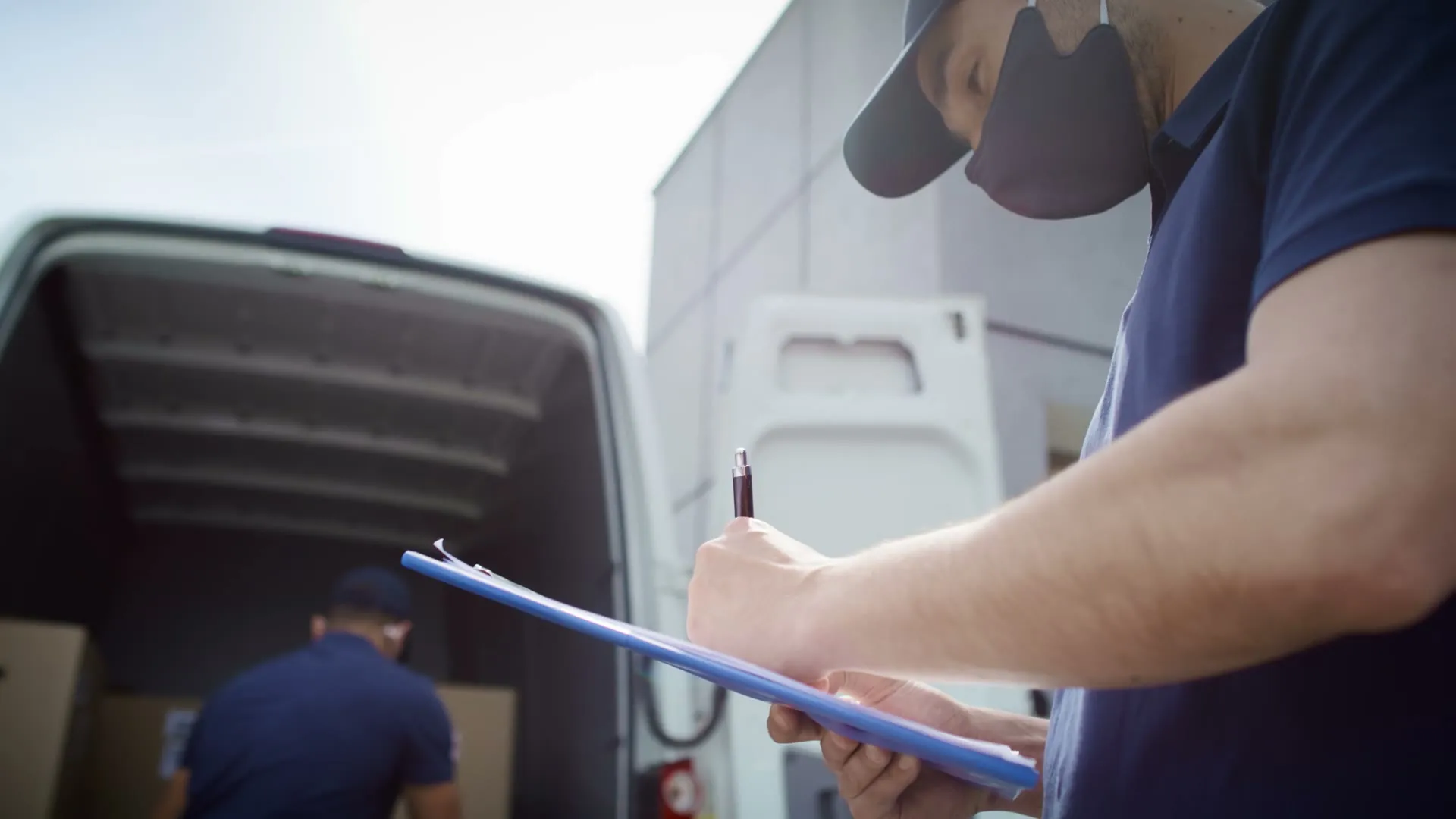 A delivery driver signs a paper form on a clipboard next to the open door of a van.
