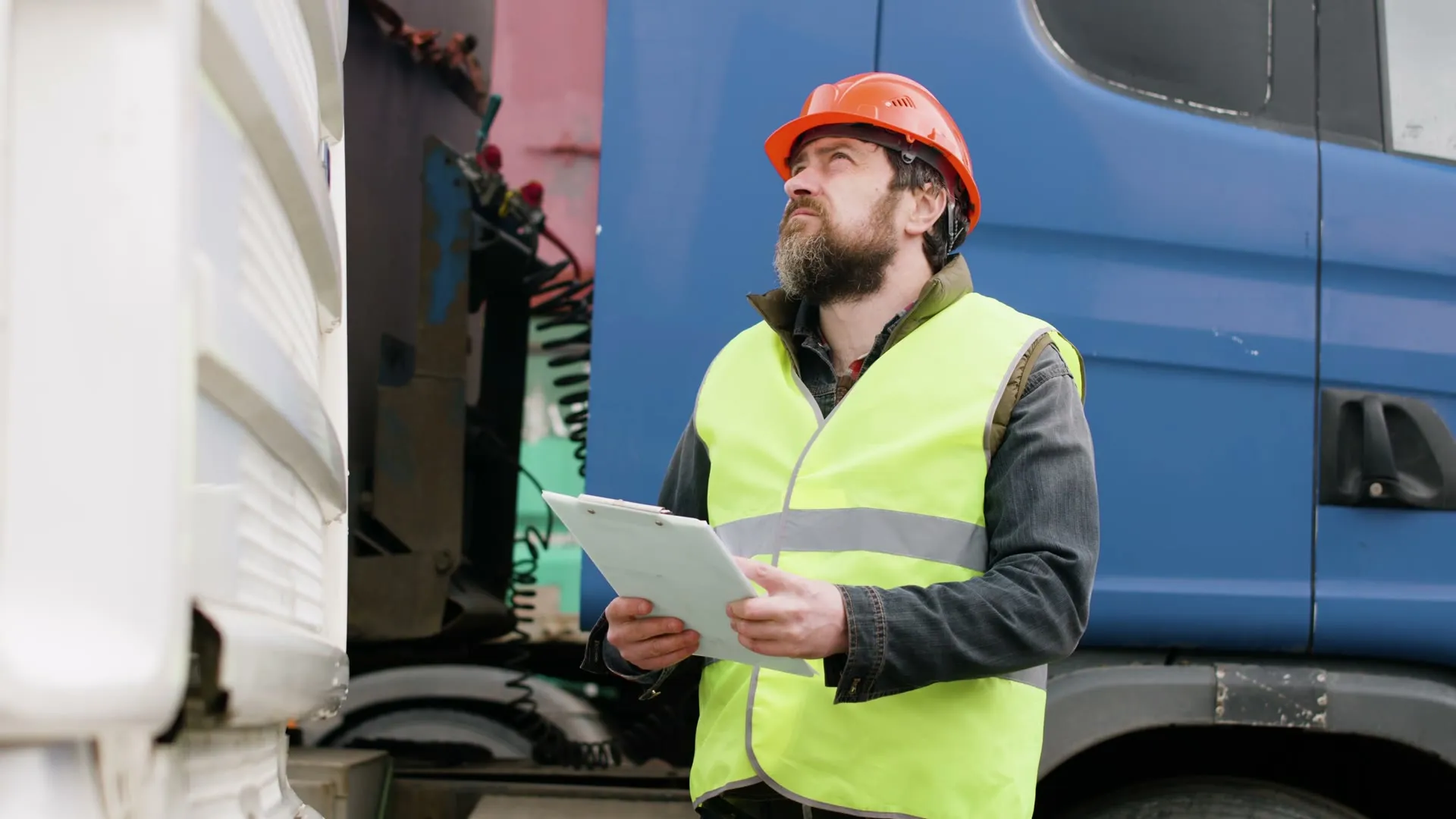A yard supervisor in a hard hat holds a clipboard next to the trucks.