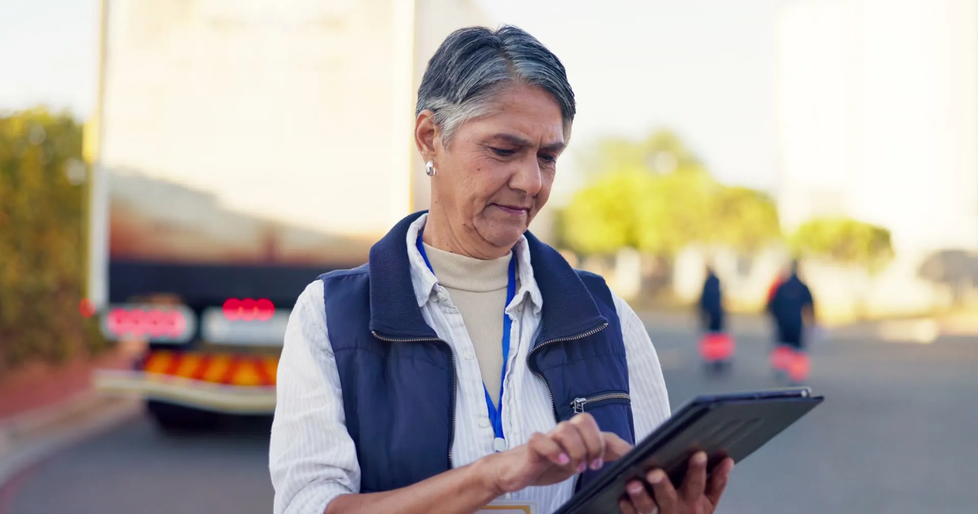 An operations manager reviews a tablet in a yard with trucks behind her.